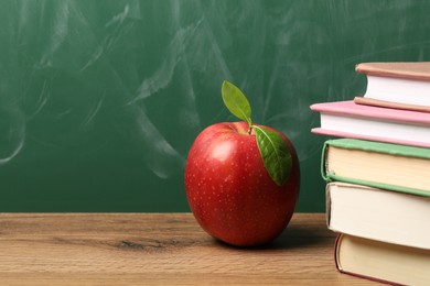 Photo of Different books and red apple on wooden table near blackboard, closeup. Space for text