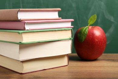 Photo of Different books and red apple on wooden table near blackboard, closeup
