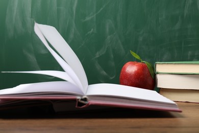 Photo of Different books and red apple on wooden table near blackboard, closeup