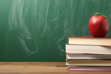 Photo of Different books and red apple on wooden table near blackboard, closeup. Space for text