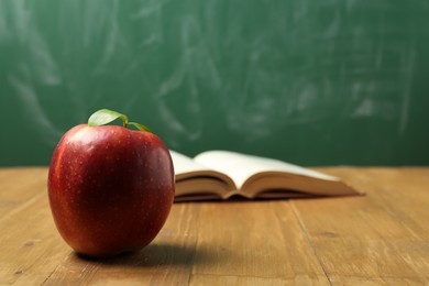 Photo of Red apple and open book on wooden table near blackboard, closeup