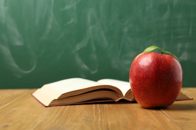 Photo of Red apple and open book on wooden table near blackboard, closeup