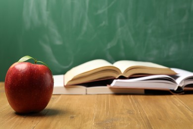 Photo of Red apple and different books on wooden table near blackboard, closeup