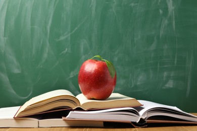 Photo of Different books and red apple on wooden table near blackboard