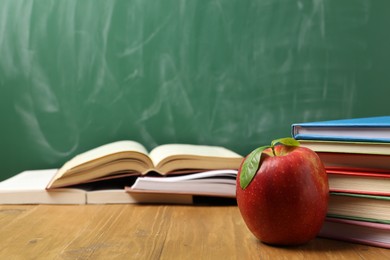 Photo of Different books and red apple on wooden table near blackboard, closeup