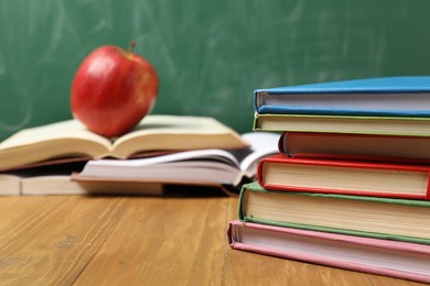 Photo of Different books and red apple on wooden table near blackboard, closeup