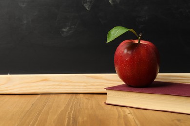 Photo of Book and red apple on wooden table near blackboard, closeup. Space for text