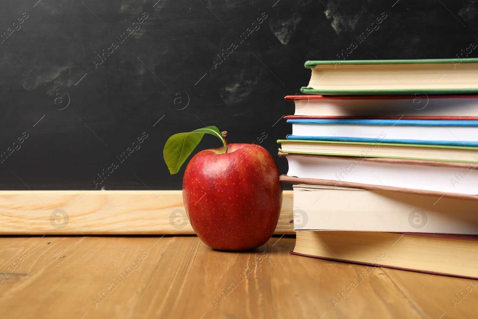 Different books and red apple on wooden table near blackboard, closeup. Space for text Photo of Different books and red apple on wooden table near blackboard, closeup. Space for text