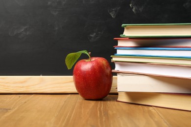 Photo of Different books and red apple on wooden table near blackboard, closeup. Space for text