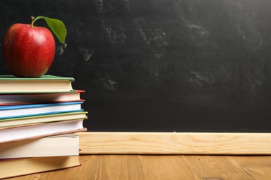 Photo of Different books and red apple on wooden table near blackboard, closeup. Space for text