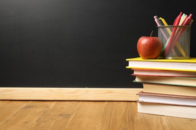 Photo of Different books, pens and red apple on wooden table near blackboard, space for text