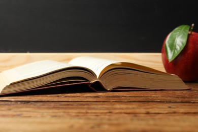 Photo of Open book and red apple on wooden table near blackboard, closeup