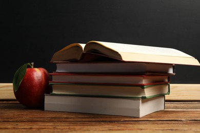Photo of Different books and red apple on wooden table near blackboard