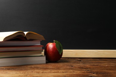 Photo of Different books and red apple on wooden table near blackboard, space for text