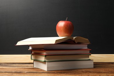 Photo of Different books and red apple on wooden table near blackboard
