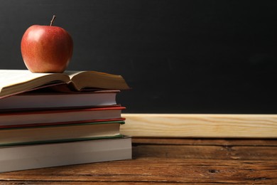 Photo of Different books and apple on wooden table near blackboard, closeup. Space for text