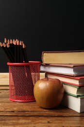Photo of Different books, pencils and apple on wooden table near blackboard, closeup