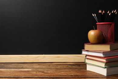 Photo of Different books, pencils and apple on wooden table near blackboard, space for text