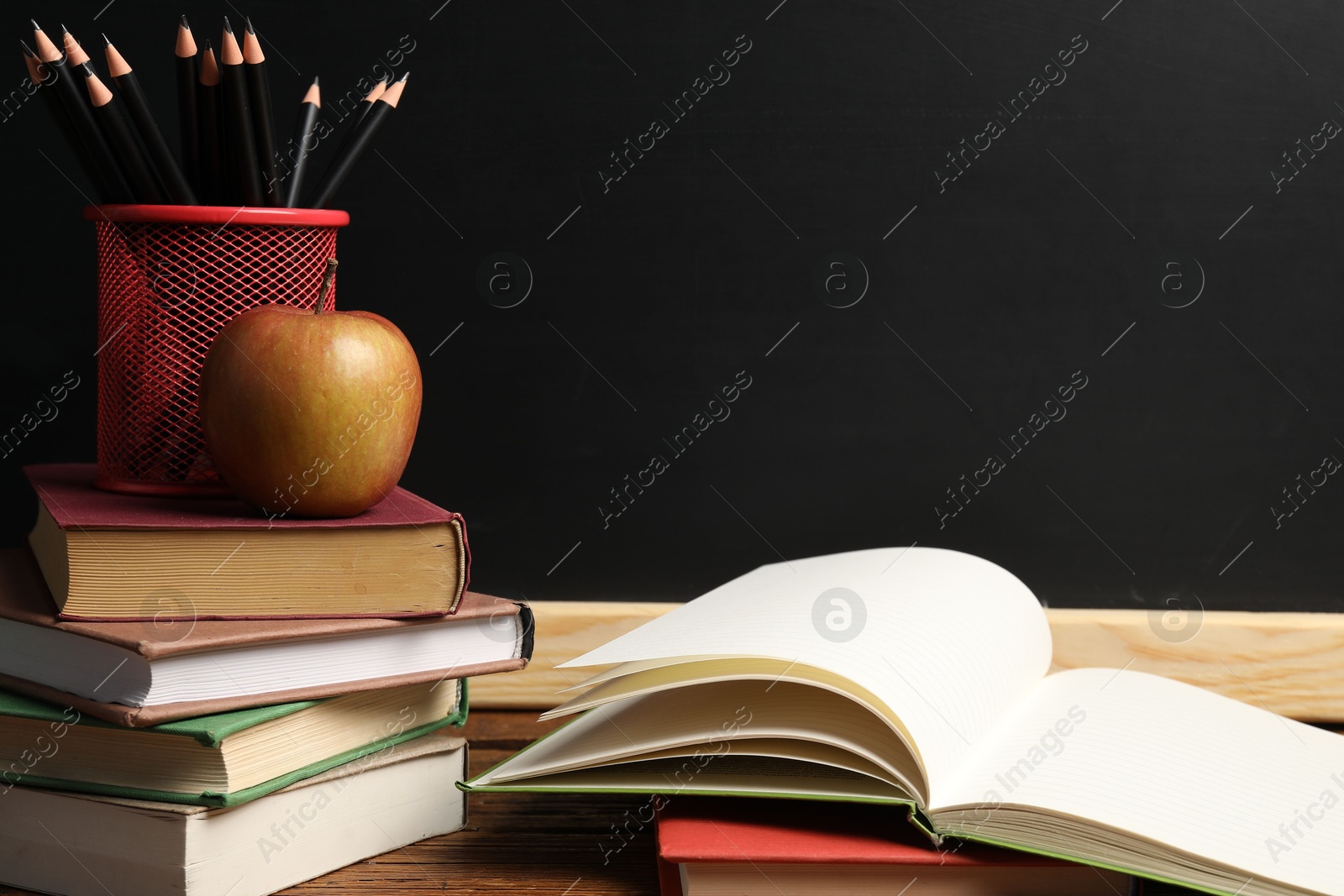 Different books, pencils and apple on wooden table near blackboard, closeup Photo of Different books, pencils and apple on wooden table near blackboard, closeup