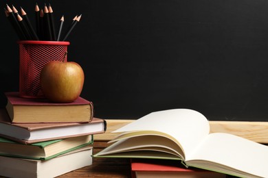 Photo of Different books, pencils and apple on wooden table near blackboard, closeup
