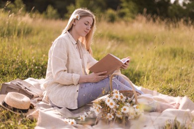 Photo of Beautiful woman with bouquet of wildflowers reading book on blanket in meadow
