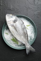 Photo of Fresh raw dourade fish, salt and rosemary in bowl on black table, top view. Organic seafood