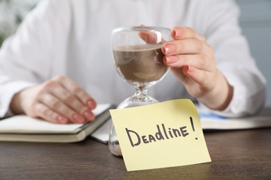 Photo of Deadline. Woman with hourglass and notebooks at wooden table in office, closeup