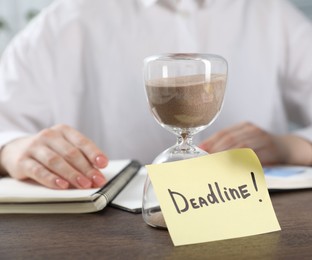 Photo of Deadline. Woman with notebooks working at wooden table in office, focus on hourglass and sticky note