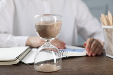Photo of Deadline. Woman with notebooks working at wooden table in office, focus on hourglass