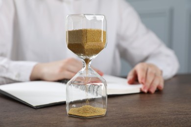Photo of Deadline. Woman with notebook working at wooden table in office, focus on hourglass