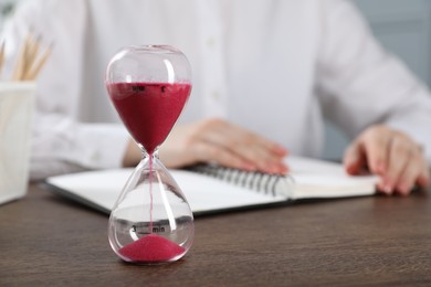 Photo of Deadline. Woman with notebook working at wooden table in office, focus on hourglass