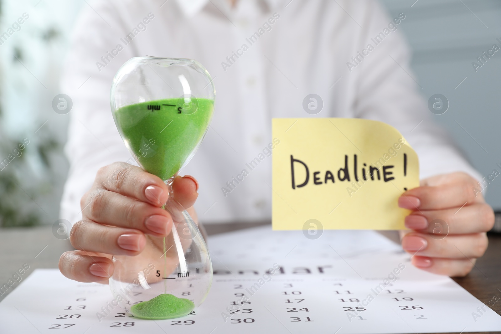 Photo of Woman holding sticky note with word Deadline and hourglass at table in office, closeup