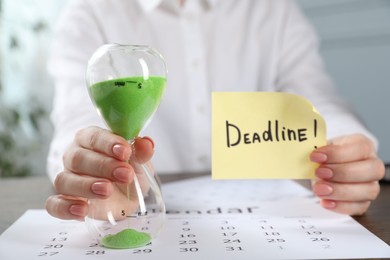 Photo of Woman holding sticky note with word Deadline and hourglass at table in office, closeup