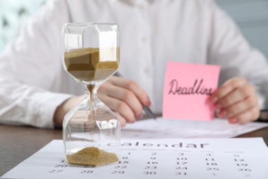 Photo of Woman holding sticky note with word Deadline while working at table in office, focus on hourglass