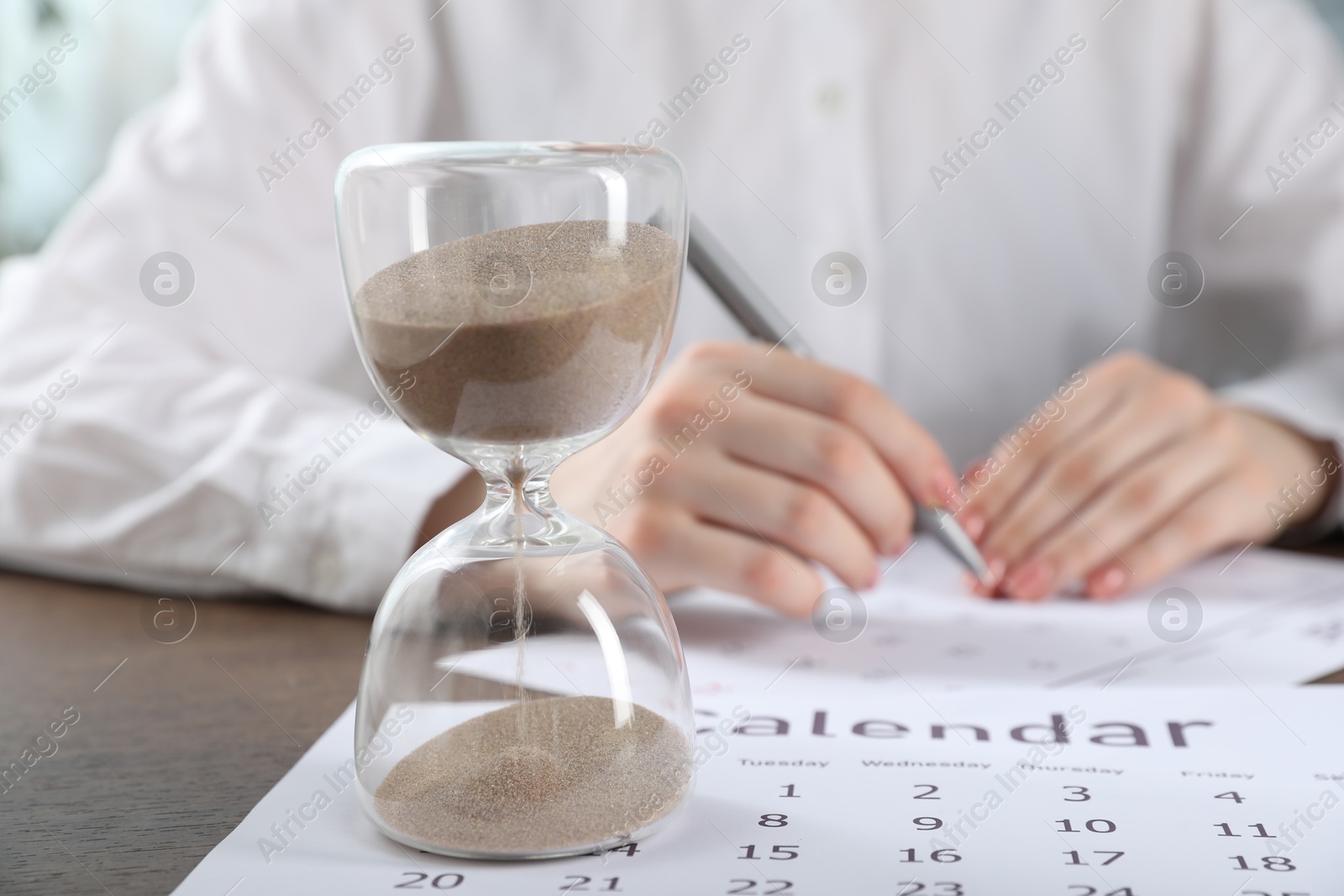 Deadline. Woman with calendar and pen at wooden table in office, focus on hourglass Photo of Deadline. Woman with calendar and pen at wooden table in office, focus on hourglass