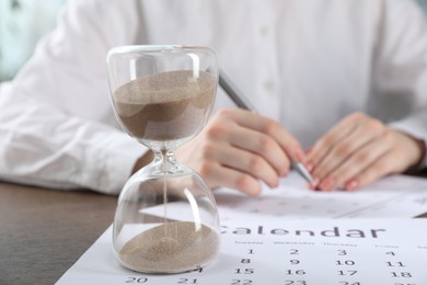 Photo of Deadline. Woman with calendar and pen at wooden table in office, focus on hourglass