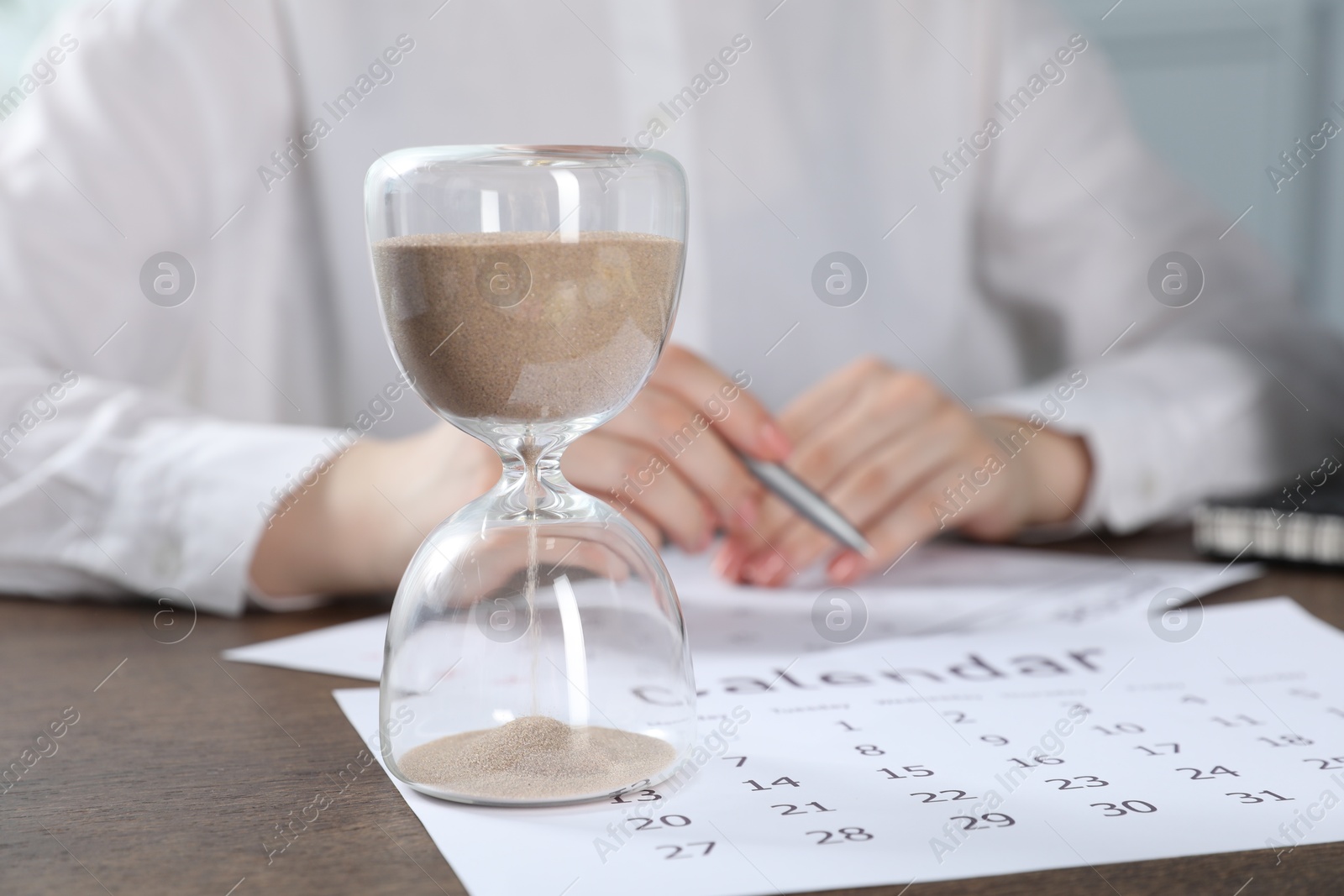 Deadline. Woman with calendar and pen at wooden table in office, focus on hourglass Photo of Deadline. Woman with calendar and pen at wooden table in office, focus on hourglass