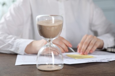 Photo of Deadline. Woman with calendar, sticky note and pen at wooden table in office, focus on hourglass