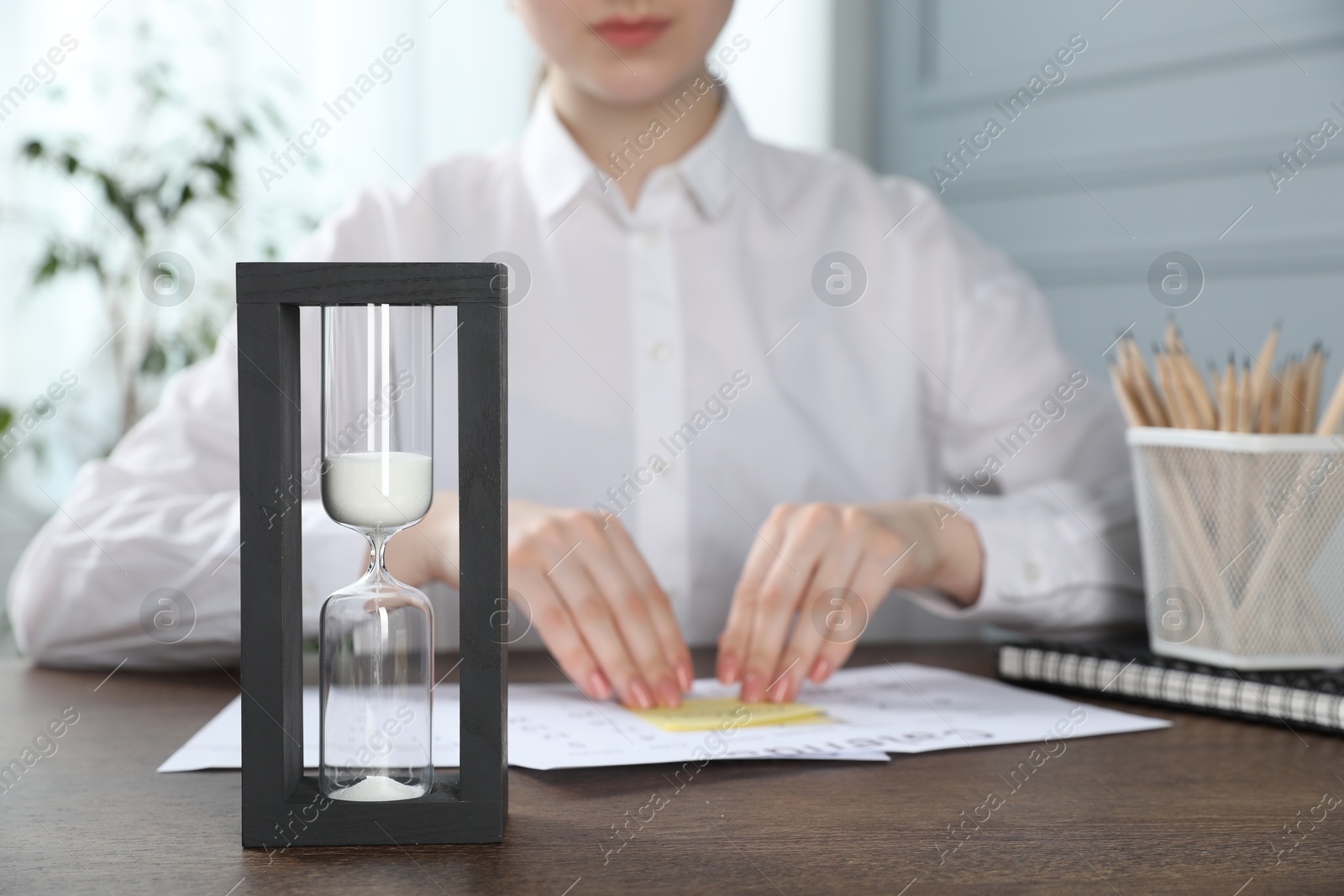 Deadline. Woman attaching sticky note to calendar at wooden table in office, focus on hourglass Photo of Deadline. Woman attaching sticky note to calendar at wooden table in office, focus on hourglass