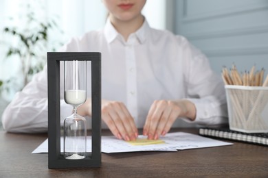 Photo of Deadline. Woman attaching sticky note to calendar at wooden table in office, focus on hourglass