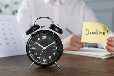 Photo of Woman holding sticky note with word Deadline while writing notes at wooden table in office, focus on alarm clock