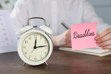 Photo of Woman holding sticky note with word Deadline while writing notes at wooden table in office, focus on alarm clock