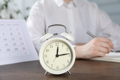 Photo of Deadline. Woman writing notes at wooden table in office, focus on alarm clock