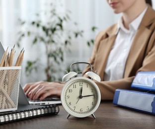 Photo of Deadline. Woman with laptop at wooden table in office, focus on alarm clock