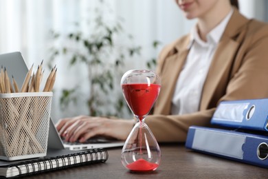 Photo of Deadline. Woman with laptop at wooden table in office, focus on hourglass