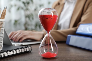Photo of Deadline. Woman with laptop at wooden table in office, focus on hourglass