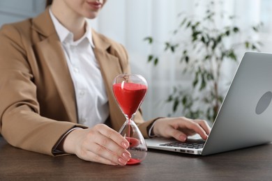 Photo of Deadline. Woman with hourglass and laptop working at wooden table in office, closeup