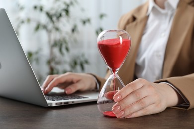 Photo of Deadline. Woman with hourglass and laptop working at wooden table in office, closeup