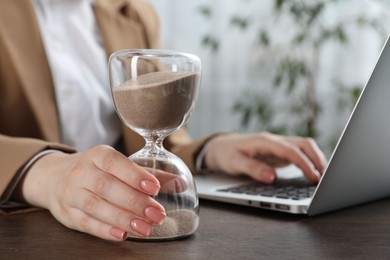 Photo of Deadline. Woman with hourglass and laptop working at wooden table in office, closeup
