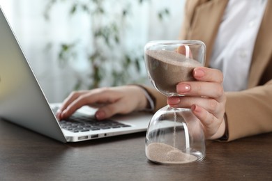 Photo of Deadline. Woman with hourglass and laptop working at wooden table in office, closeup